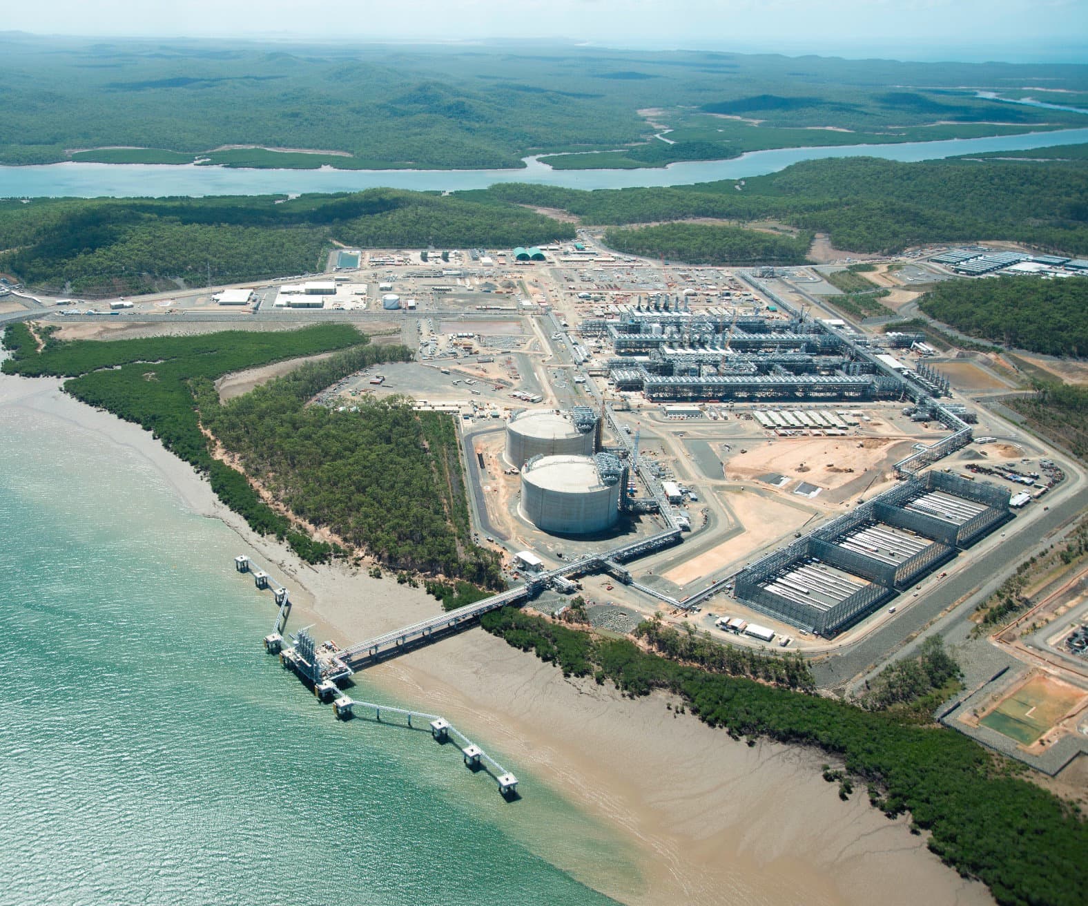 Aerial of the LNG facility hosting the Battery Energy Storage System showing the large white storage tank, industrial processing units, jetty extending into the bay, and mangrove-fringed coastline